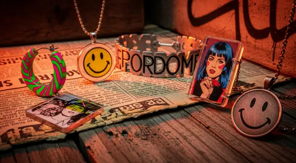 Joias Pop Art dispostas sobre um muro com grafite e jornais antigos. Inclui pingentes com rostos sorridentes em resina e broches com imagens serigrafadas, iluminados por luz neon vibrante.